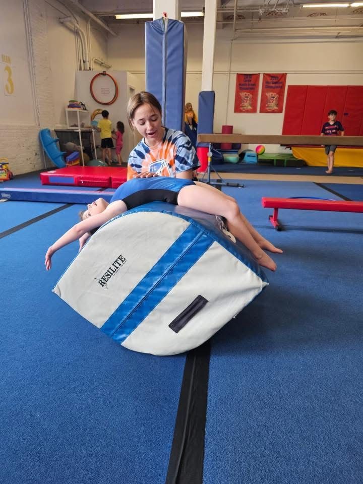 Gymnast on a training block, stretching back with assistance from a coach in a gym. Blue mats, colorful equipment visible.