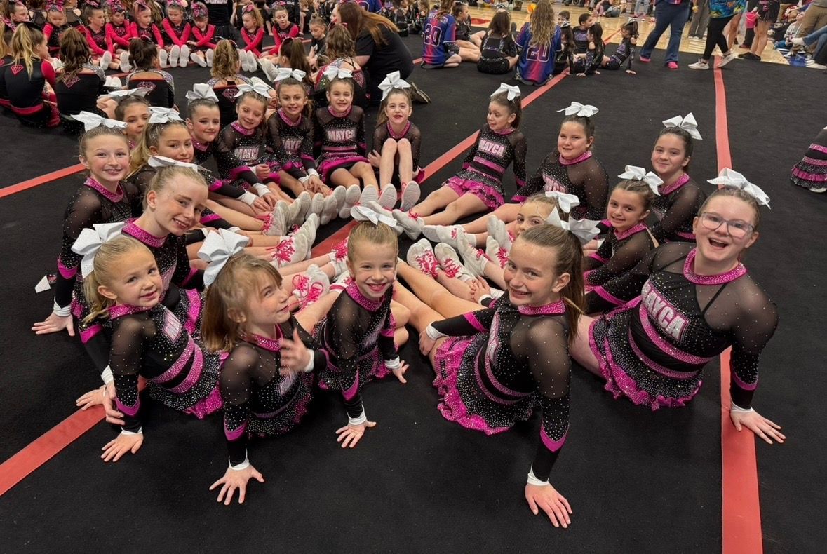 Cheerleaders in black and pink uniforms, sitting in a circle on a gym floor, smiling and posing.