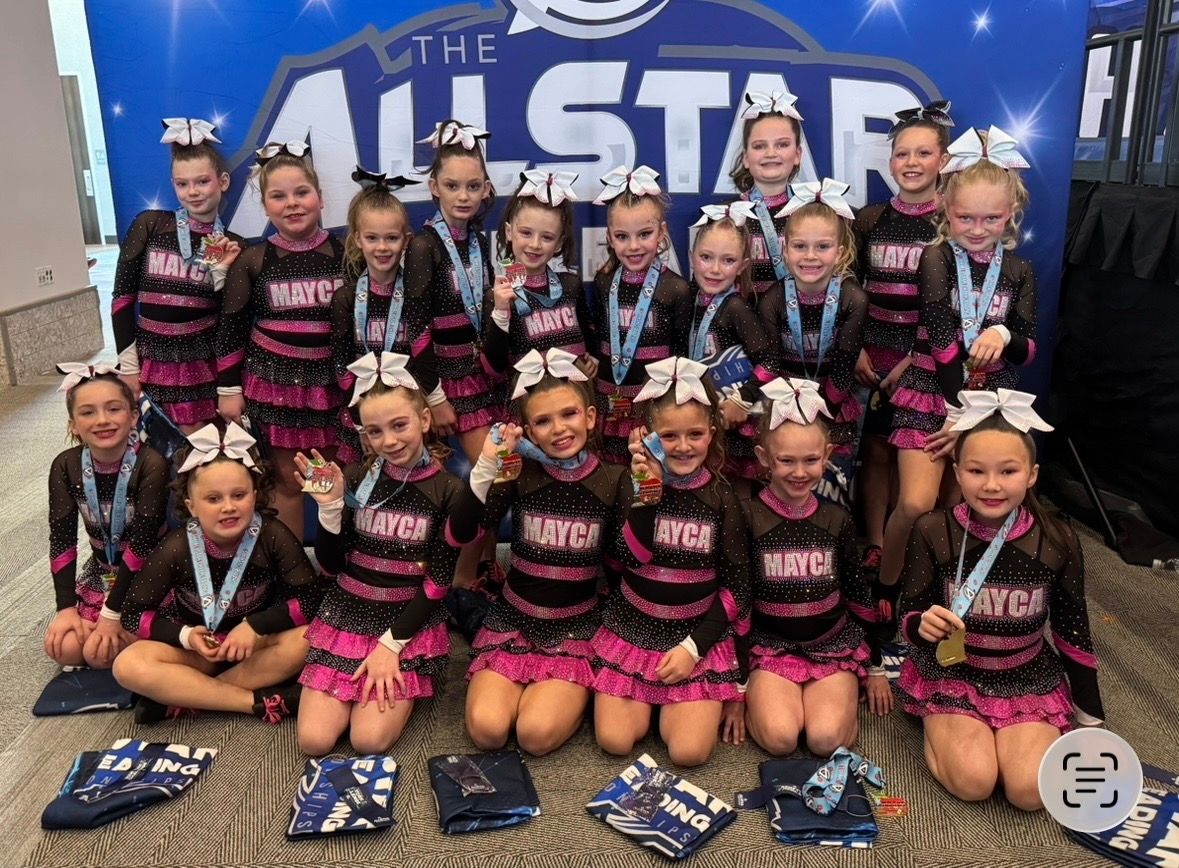 Cheerleading team posing with medals; wearing black & pink uniforms; in front of a backdrop with 