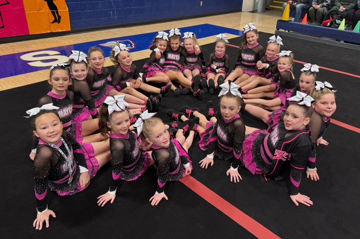 Cheerleaders in black and pink uniforms, grouped in a circle on a mat, smiling.