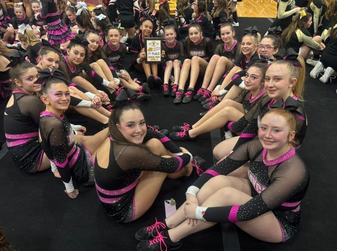 Cheerleading team in black and pink uniforms, seated in a circle, holding a trophy on a mat.
