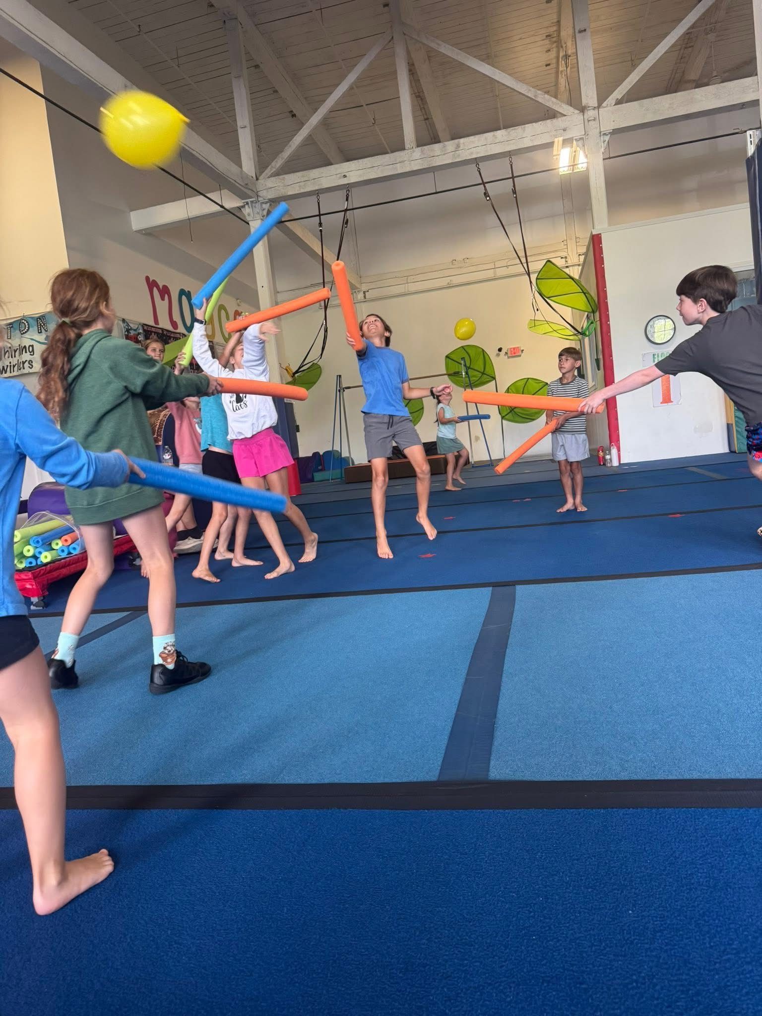 Kids playing with pool noodles and balloons in a gymnasium, some are holding the noodles like swords.