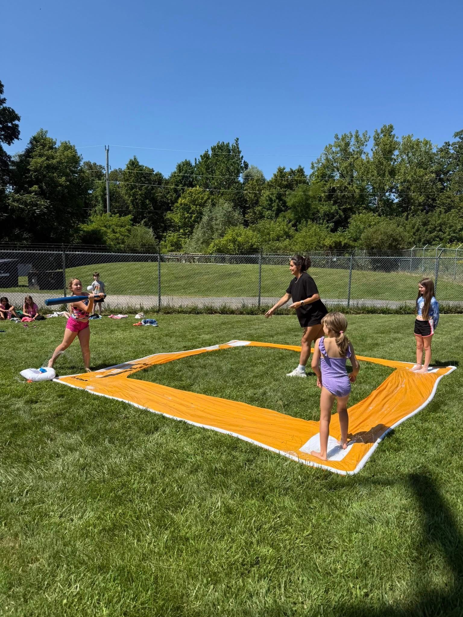 Children playing baseball on a grass field, outlined by yellow towels. Sunny day.