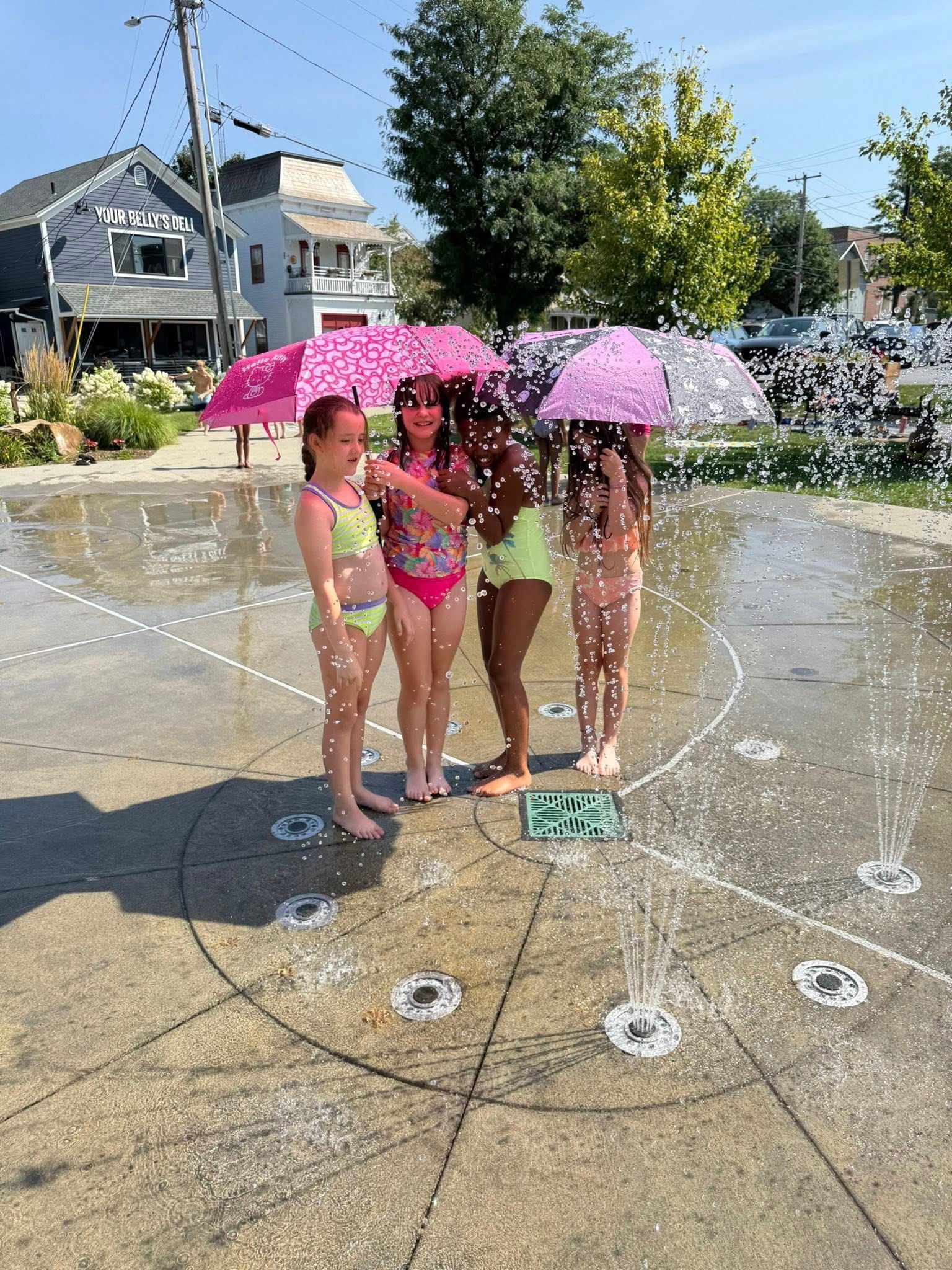 Four people in swimsuits under pink umbrellas, playing in a splash pad.