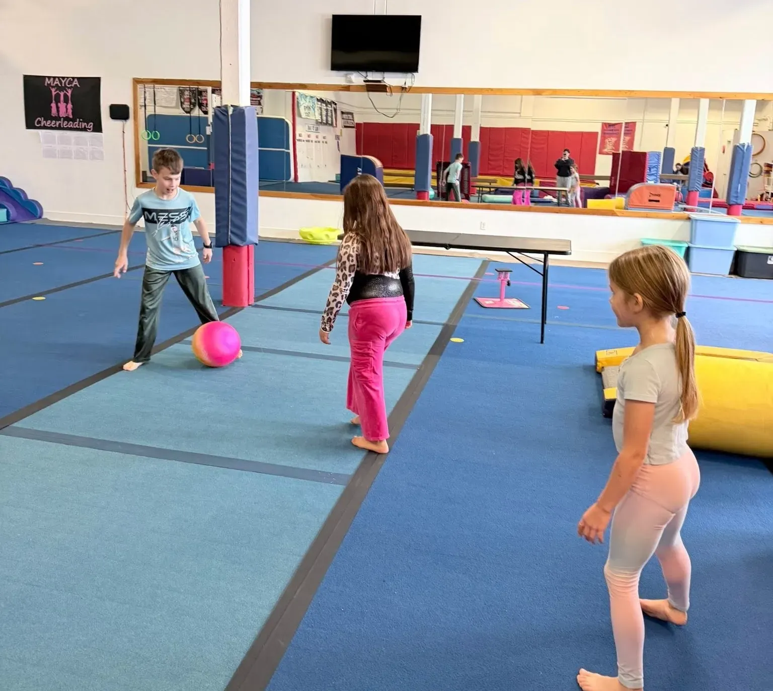 Three children playing with a pink ball on a blue gym floor.