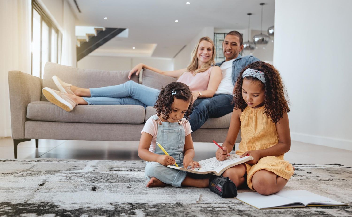 Family relaxing: parents on a couch, two young children drawing on the floor. Modern living room setting.