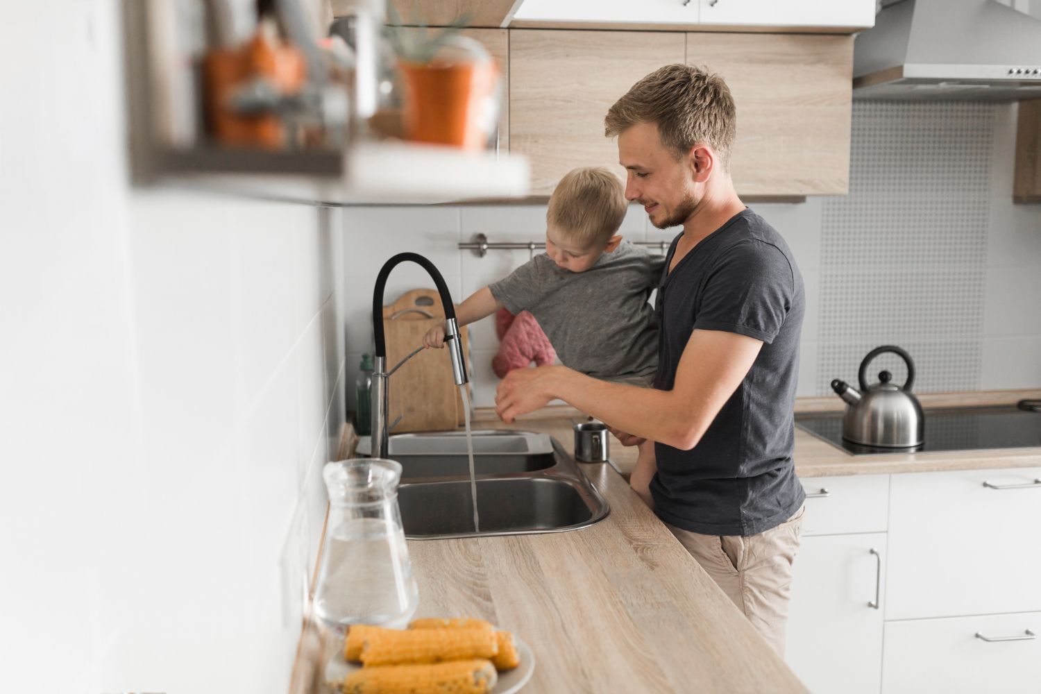 Man holding child at a kitchen sink, washing hands. Light wood counters.