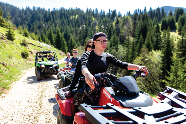 People riding ATVs on a dirt road through a forested mountain area.