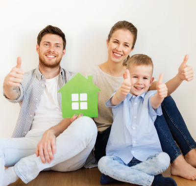 Family smiles and gives thumbs-up, holding a green house cutout. Sitting against a white wall.