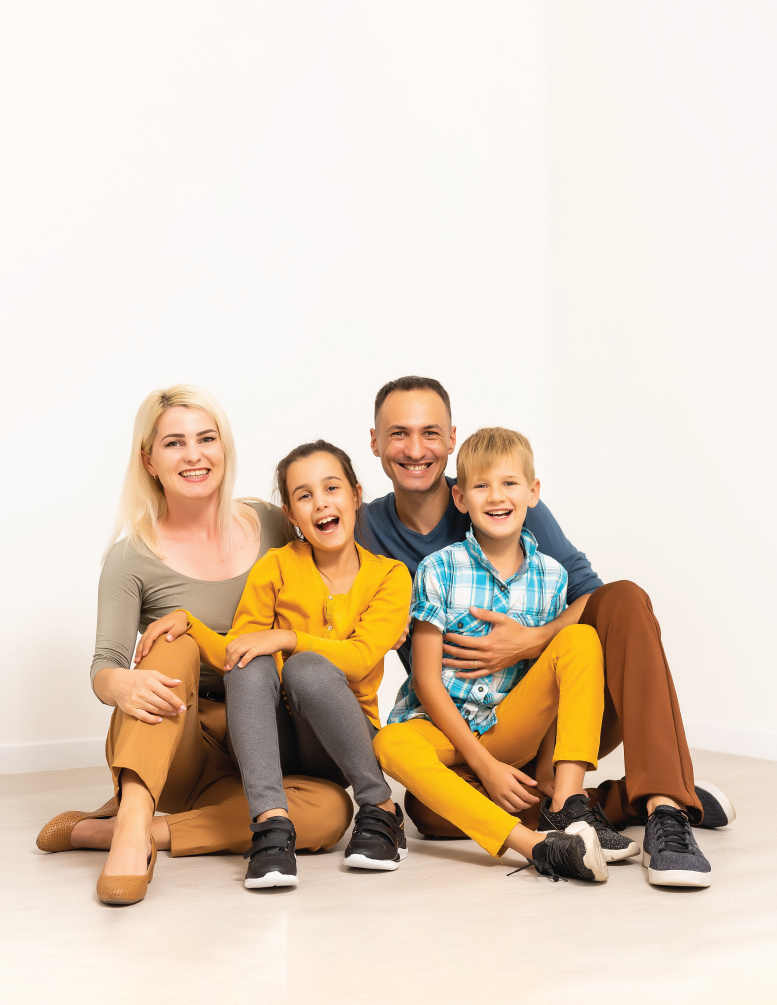 Family sitting on floor, smiling. Woman in brown, girl in yellow, man in blue, boy in blue/yellow. White background.