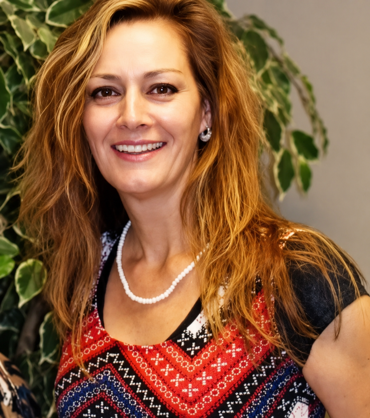 A woman wearing a necklace and earrings smiles in front of a plant