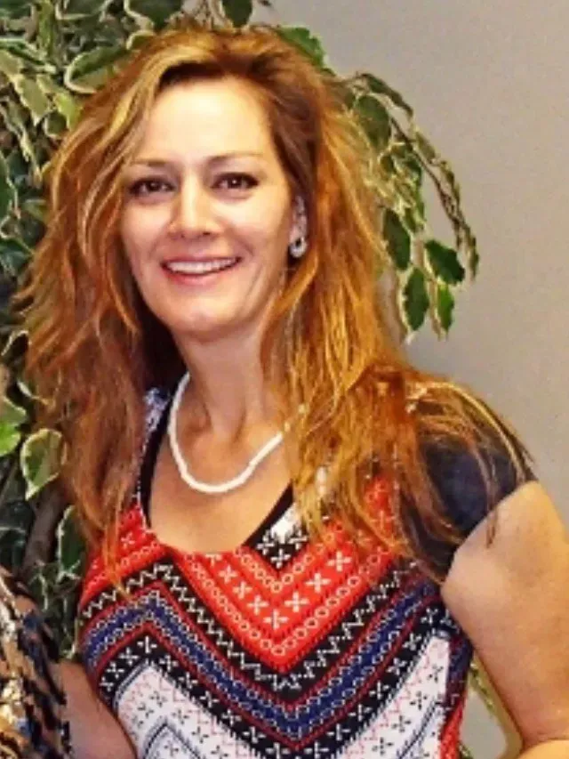 A woman wearing a necklace and earrings smiles in front of a plant