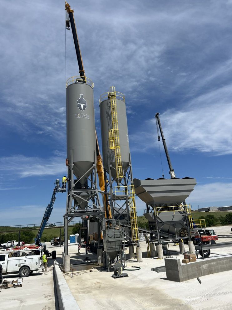 Construction site with three large grey silos and crane, under a blue sky with clouds.