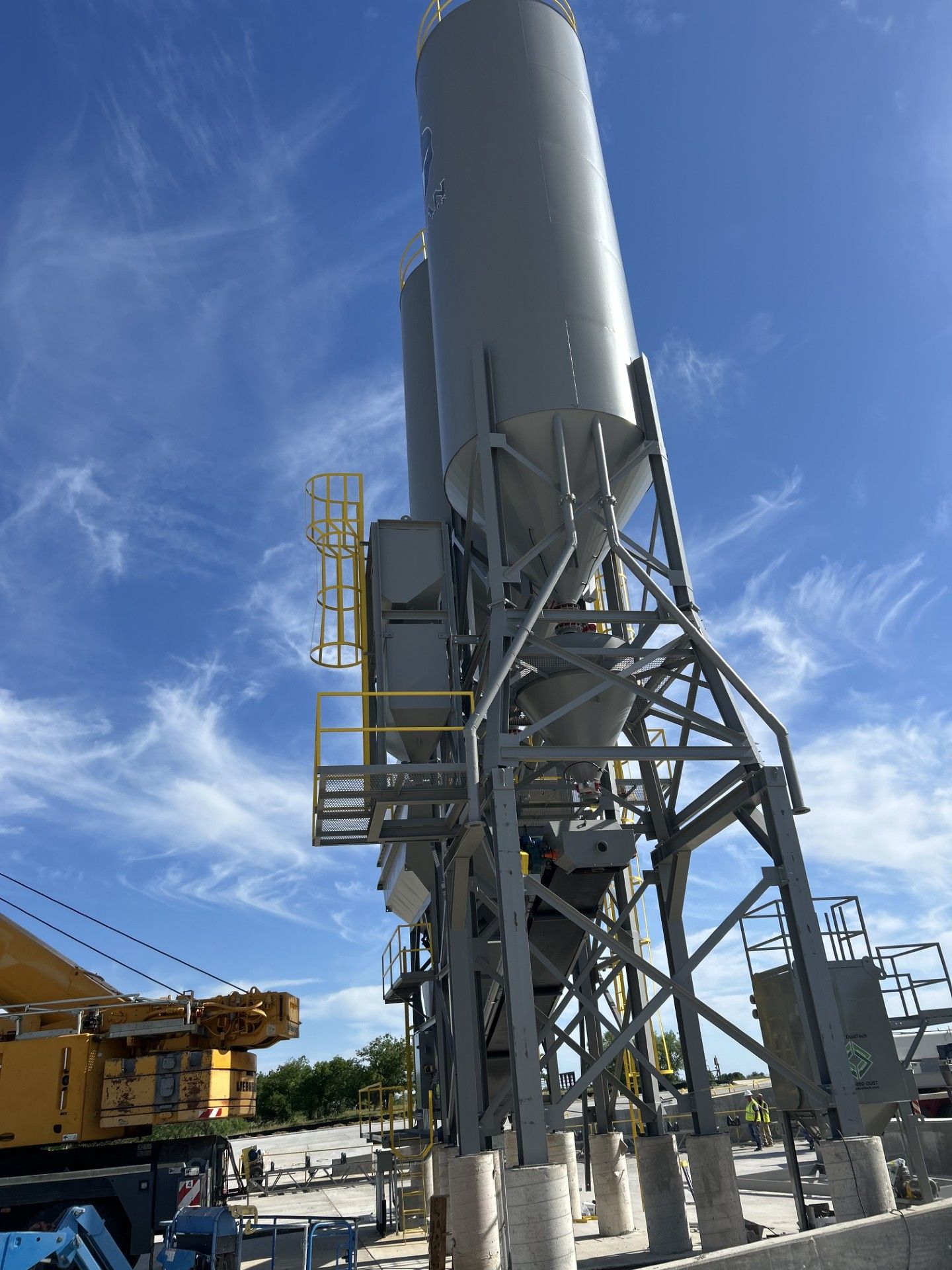 Gray industrial silos against a blue sky, supported by steel scaffolding, yellow safety features.