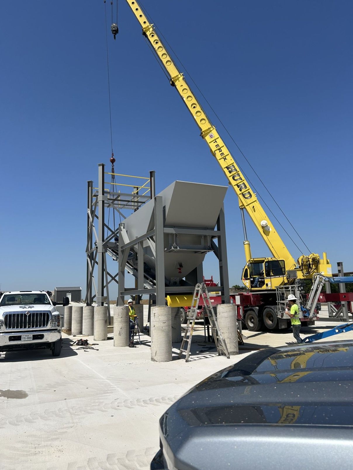 Construction site: crane lifting machinery framework, workers in hard hats, concrete pillars. Blue sky.