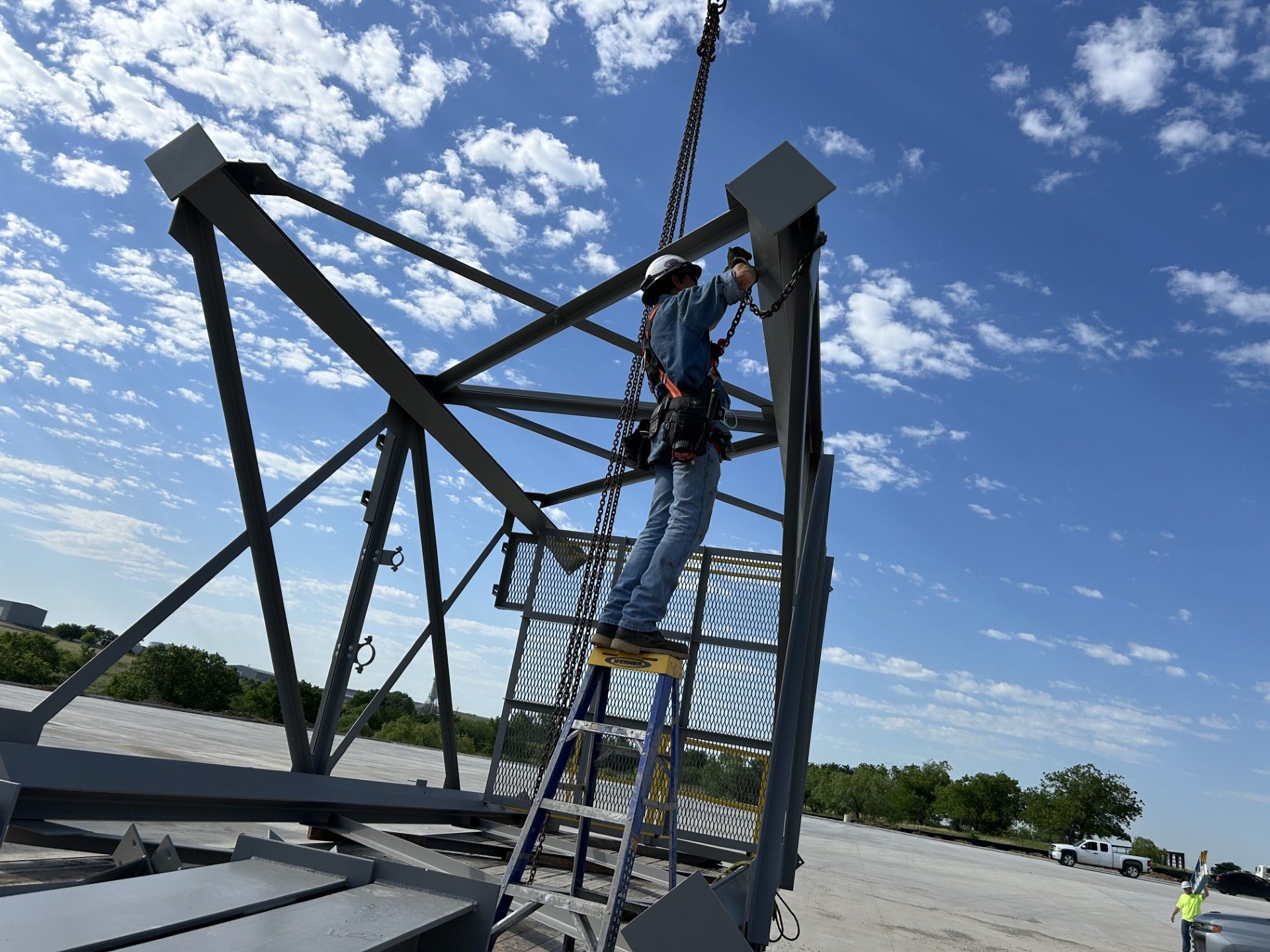 Worker on ladder attached to a metal structure, secured by a harness, under a blue sky.