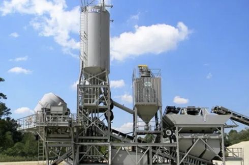 Concrete mixing plant against a blue sky with clouds. Metal towers and conveyor belts are visible.