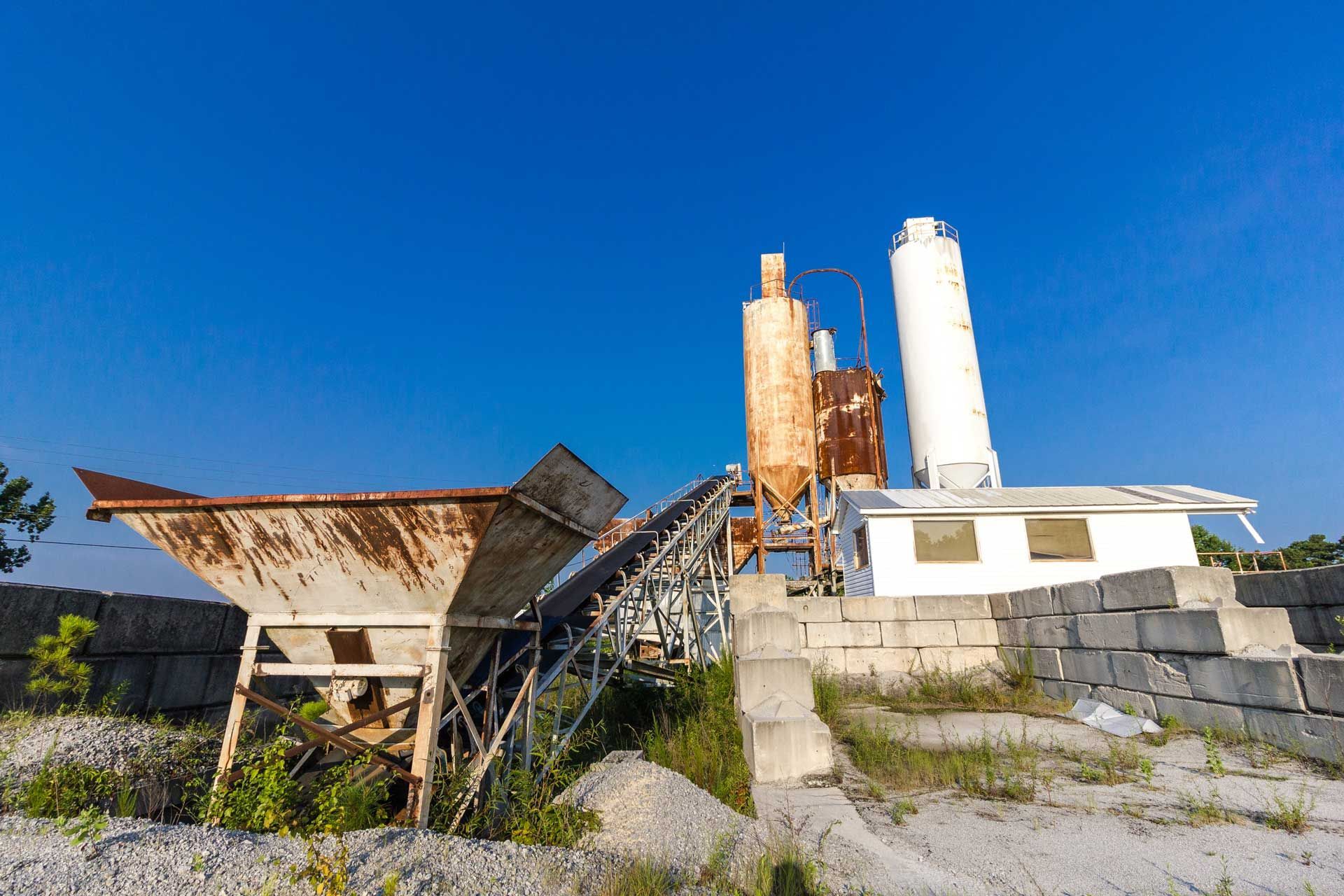 An old, rusty concrete plant with storage silos and a conveyor belt against a clear blue sky.