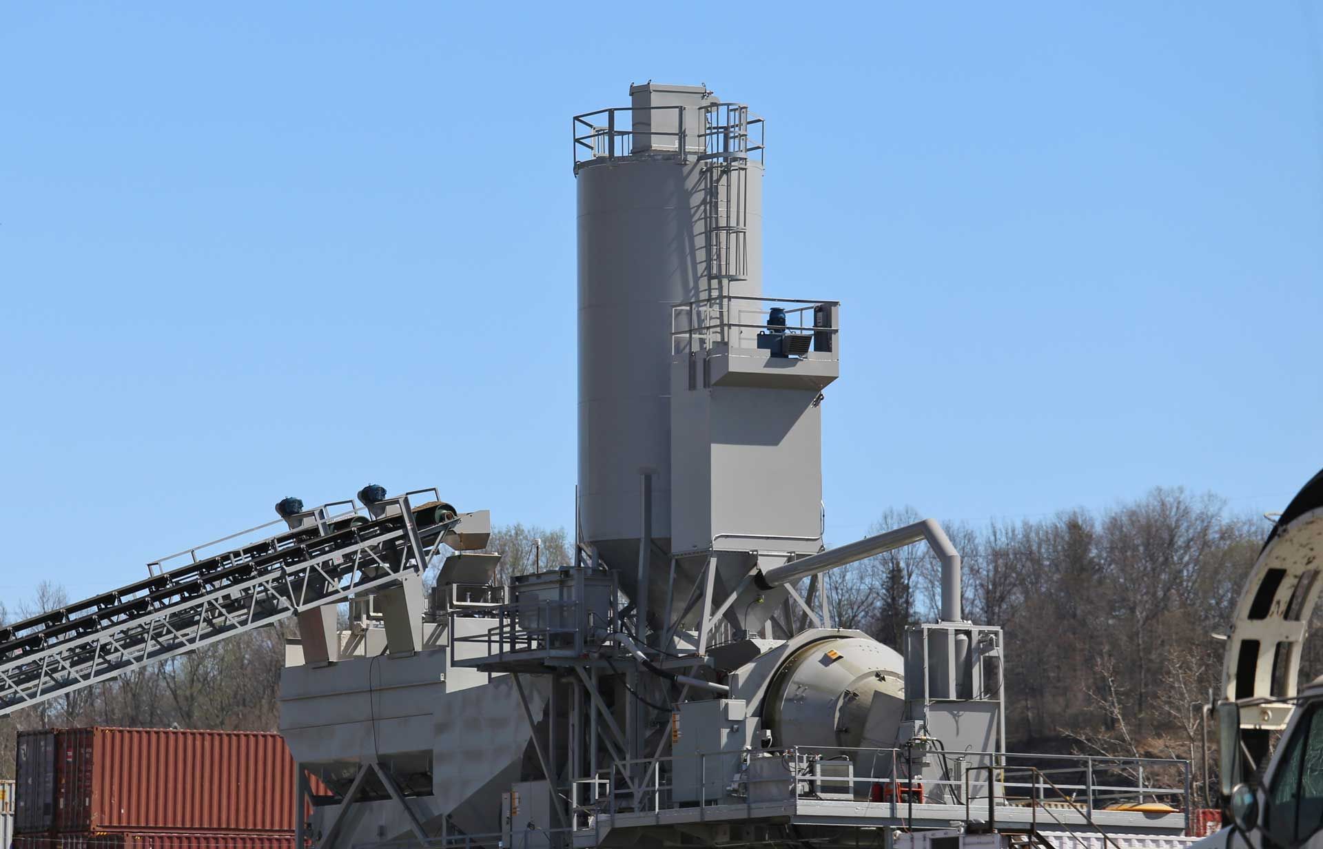 Concrete mixing plant with tall silo, conveyor belt, and mixer against a blue sky.
