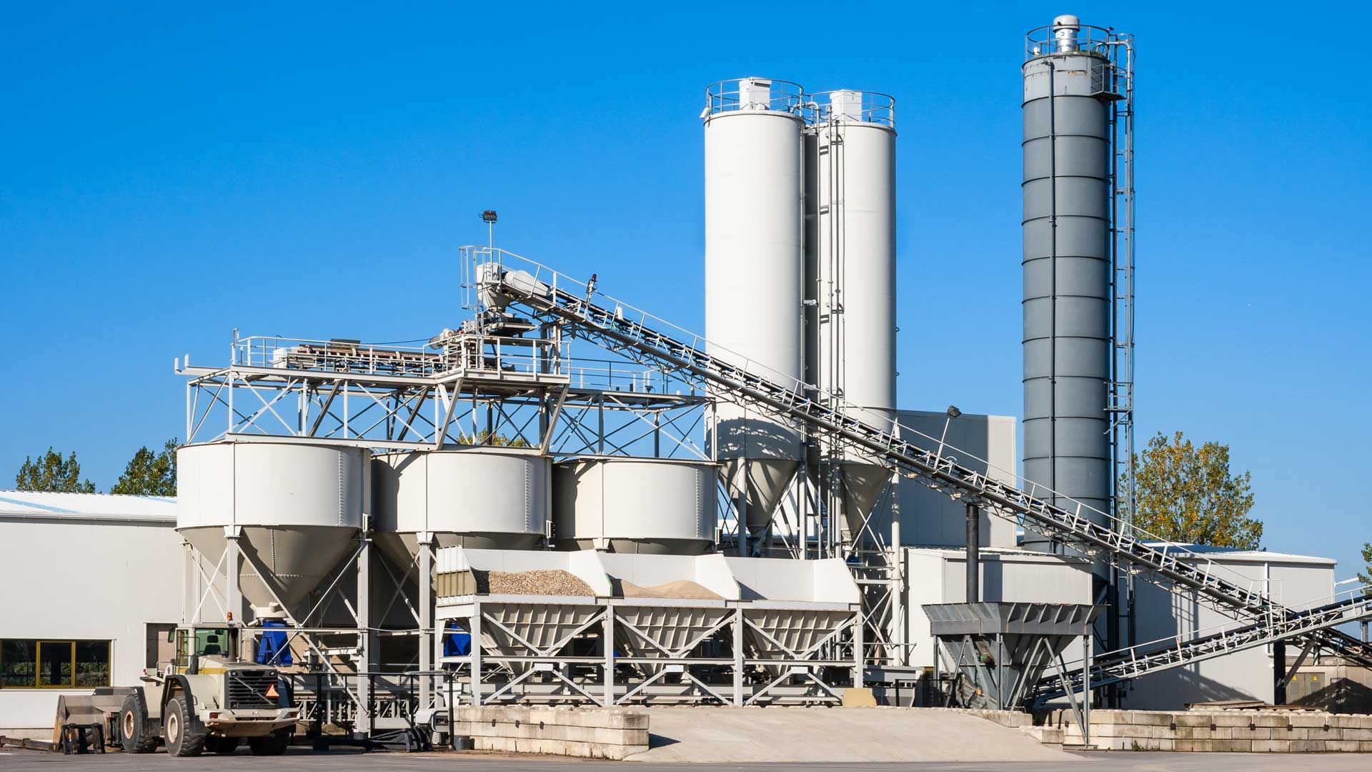 Concrete batch plant with silos, conveyor belts, and a construction vehicle under a clear blue sky.