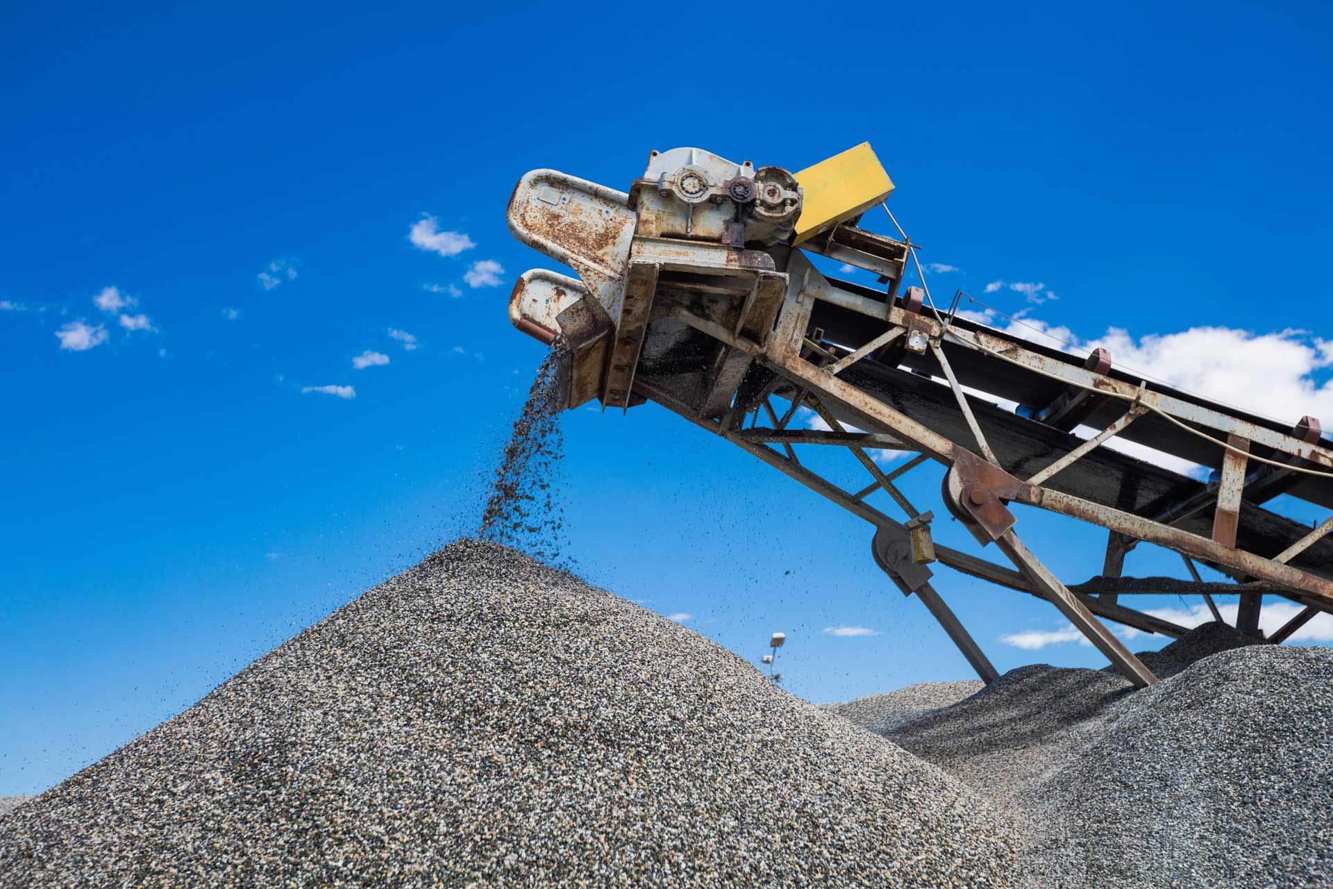A conveyor belt dispensing gravel onto a large pile under a blue sky.