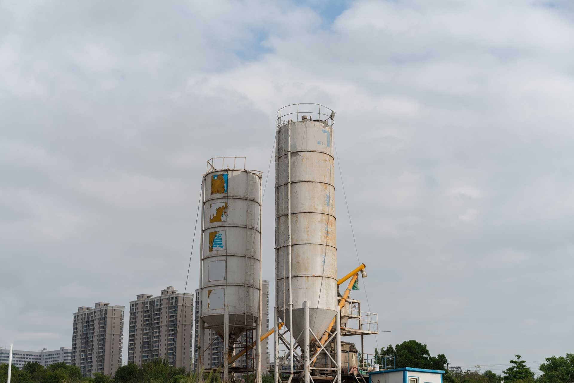 Two tall, cylindrical concrete silos against a cloudy sky, with apartment buildings in the background.