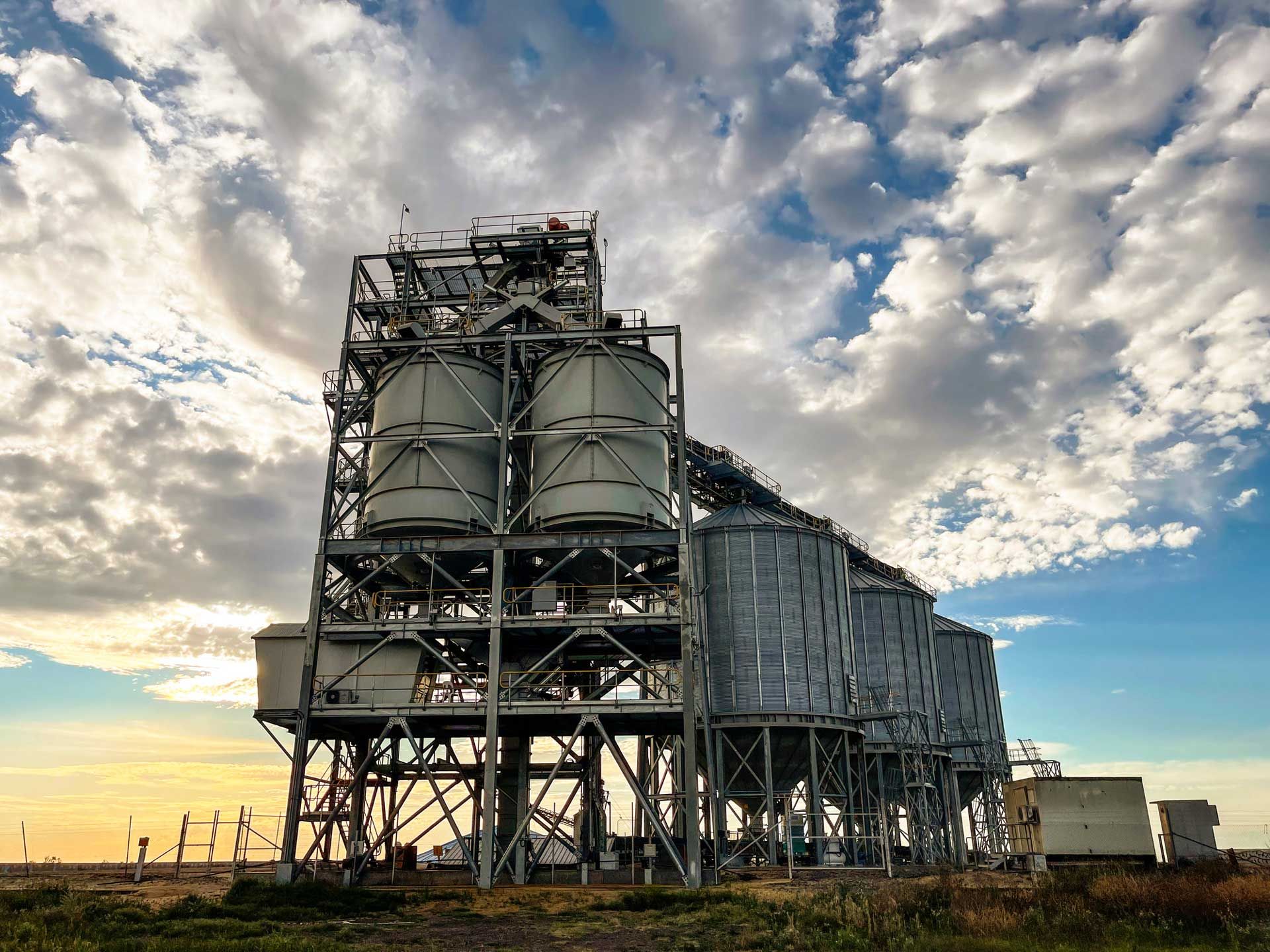 Industrial grain storage facility under a cloudy sky. Metal silos and support structure on a grassy field.