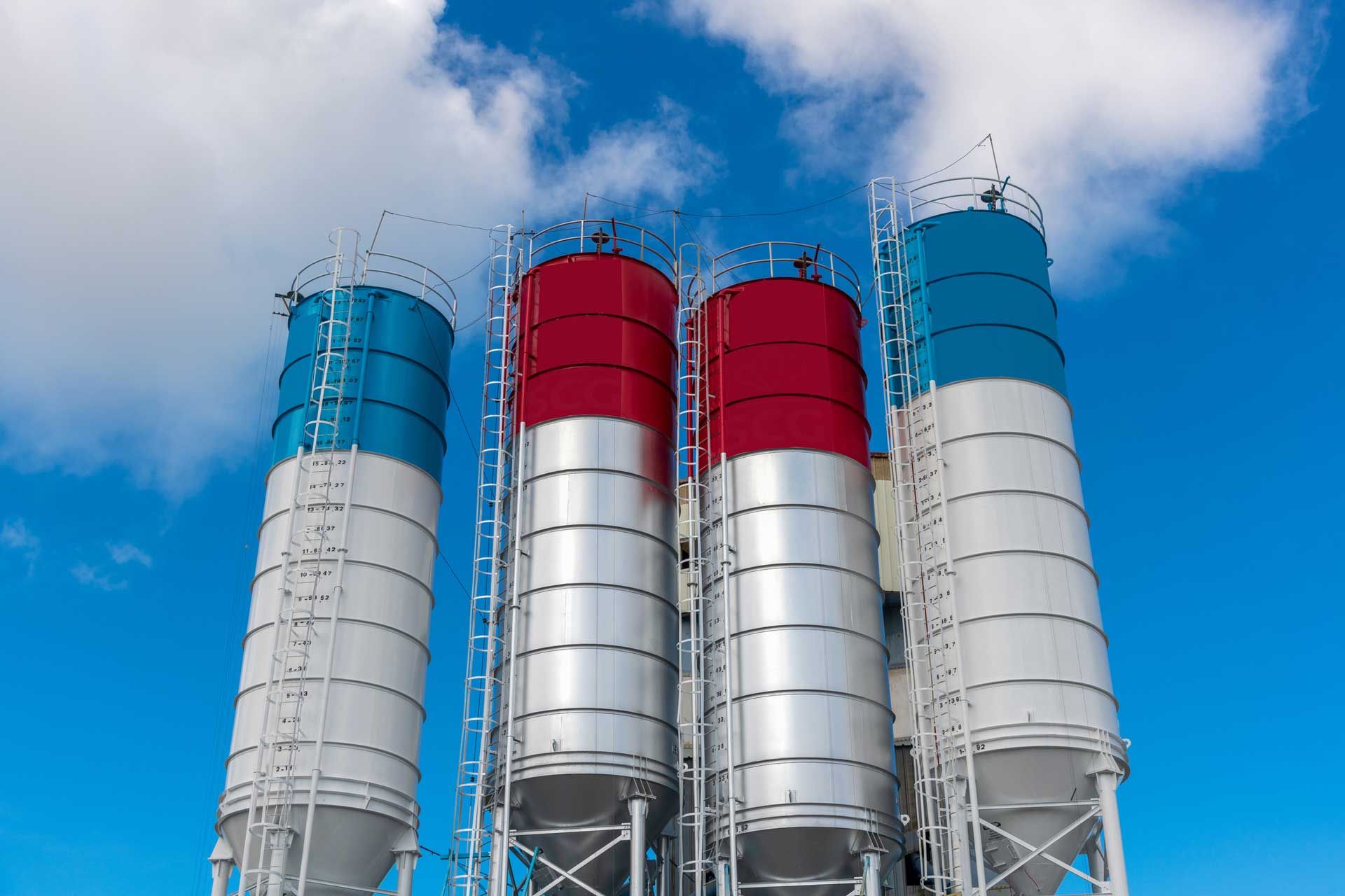 Four cylindrical industrial silos with blue, red, and white tops against a blue sky with clouds.
