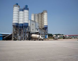 Cement mixing plant with silos, a mixer truck, and other trucks under a blue sky.