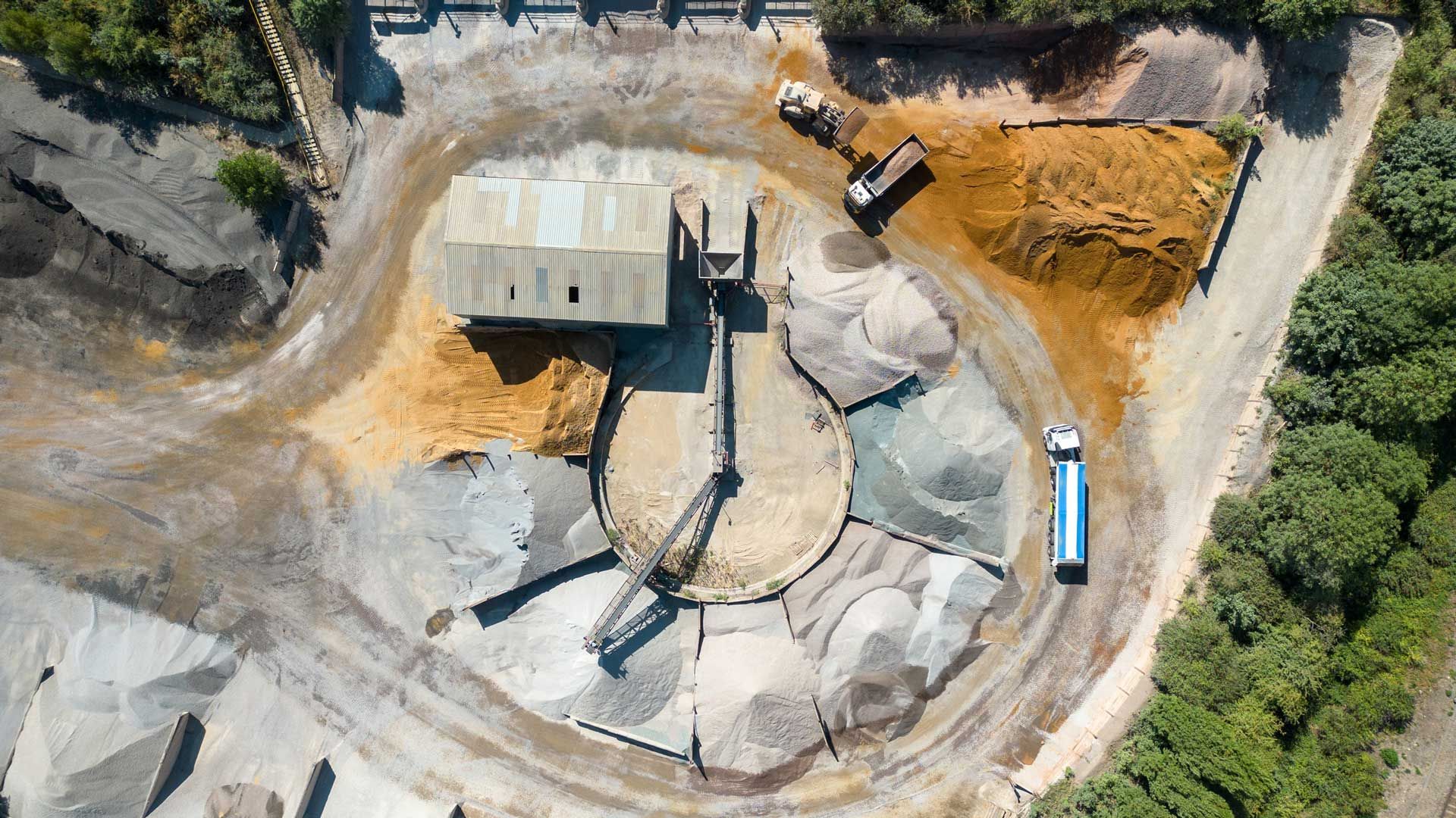 Aerial view of a quarry operation. A building, piles of material, and trucks surround a central processing area.