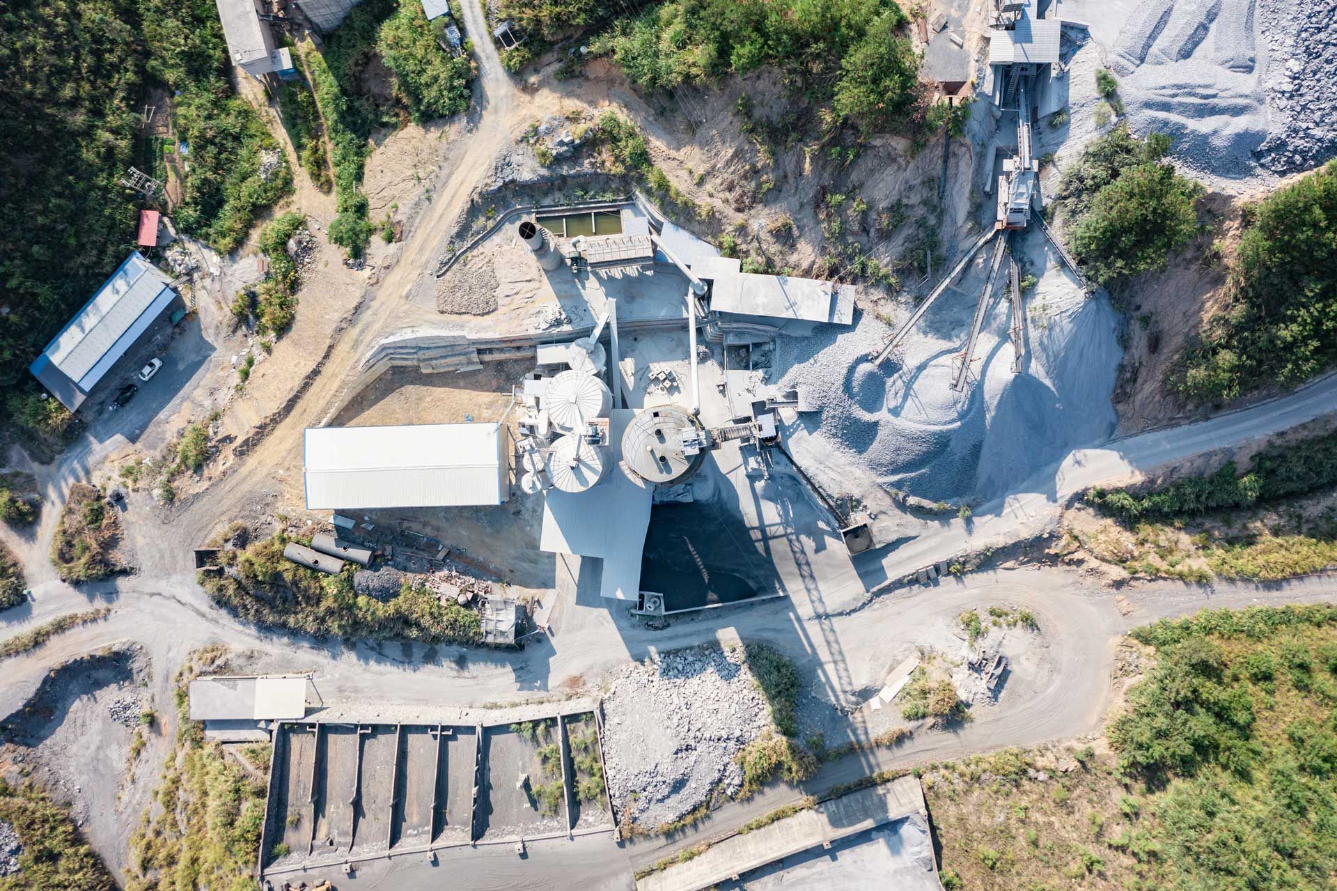 Aerial view of a quarry processing plant with machinery, silos, and piles of gray gravel surrounded by green vegetation.