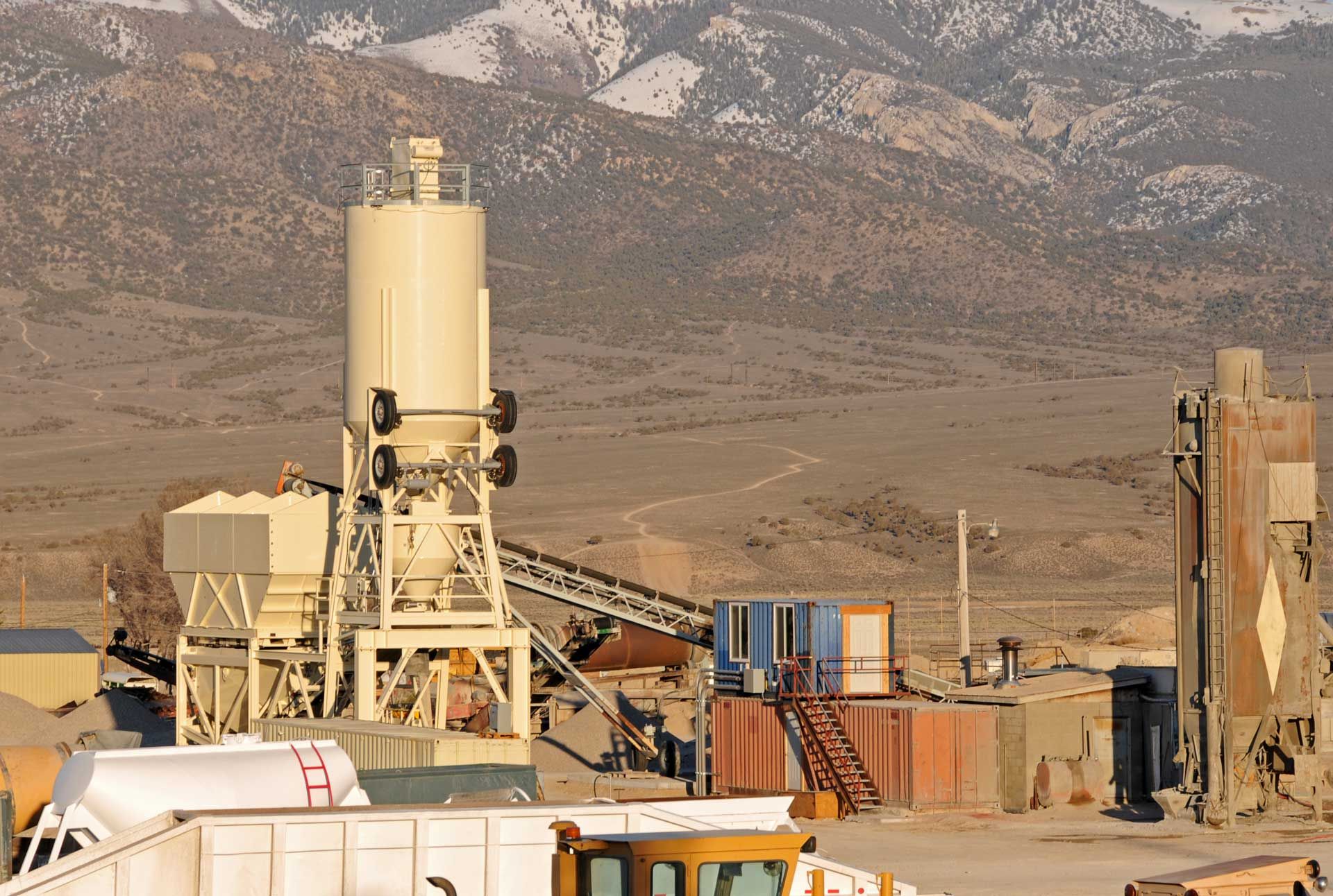 Industrial plant with tall silo, conveyor belt, and container buildings, mountains in the background.
