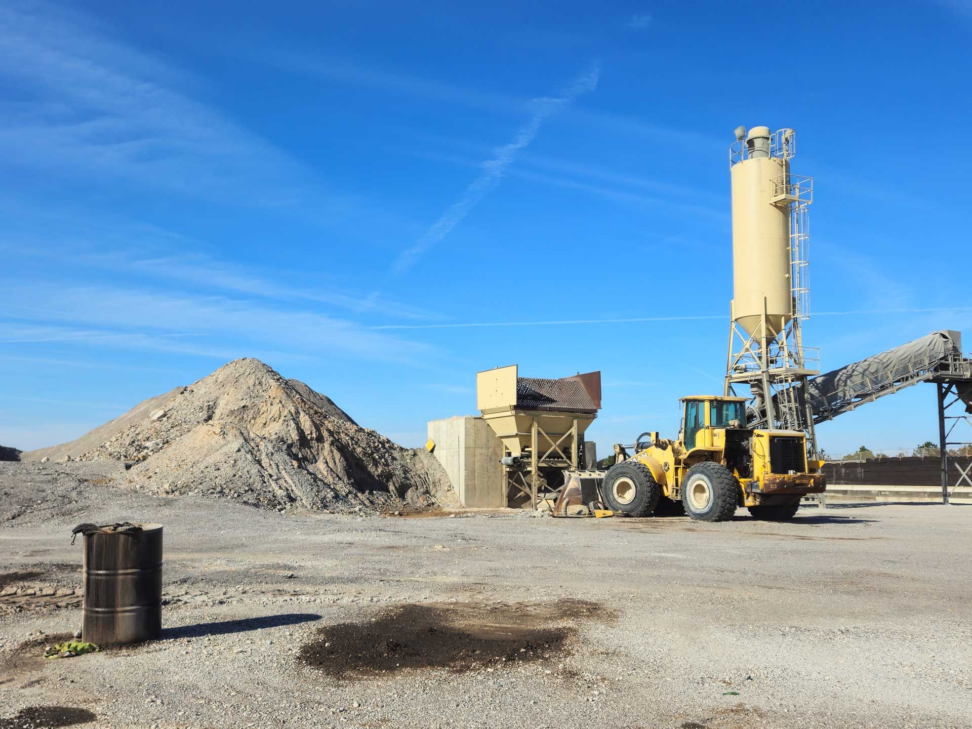 Yellow construction equipment near an industrial site. Piles of gravel and silos under a blue sky.