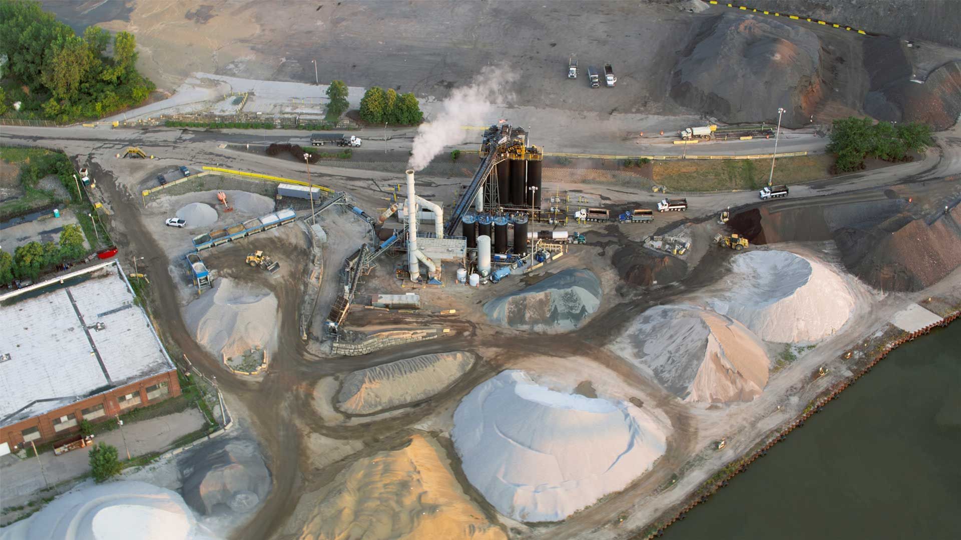 Aerial view of an industrial plant with smokestack emitting smoke, surrounded by piles of gravel and construction materials.