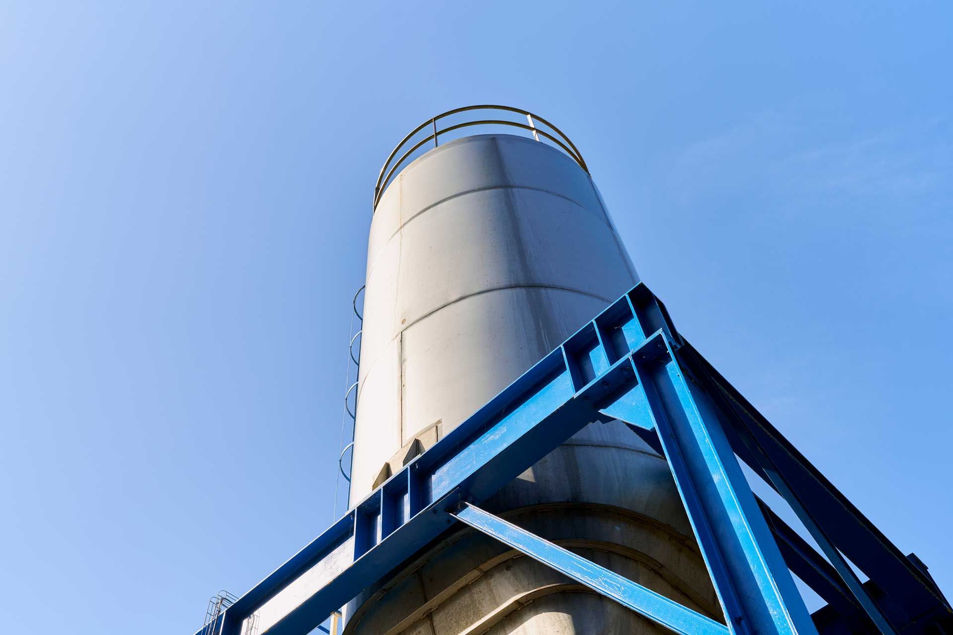Gray industrial silo with a blue metal support frame against a clear, blue sky.