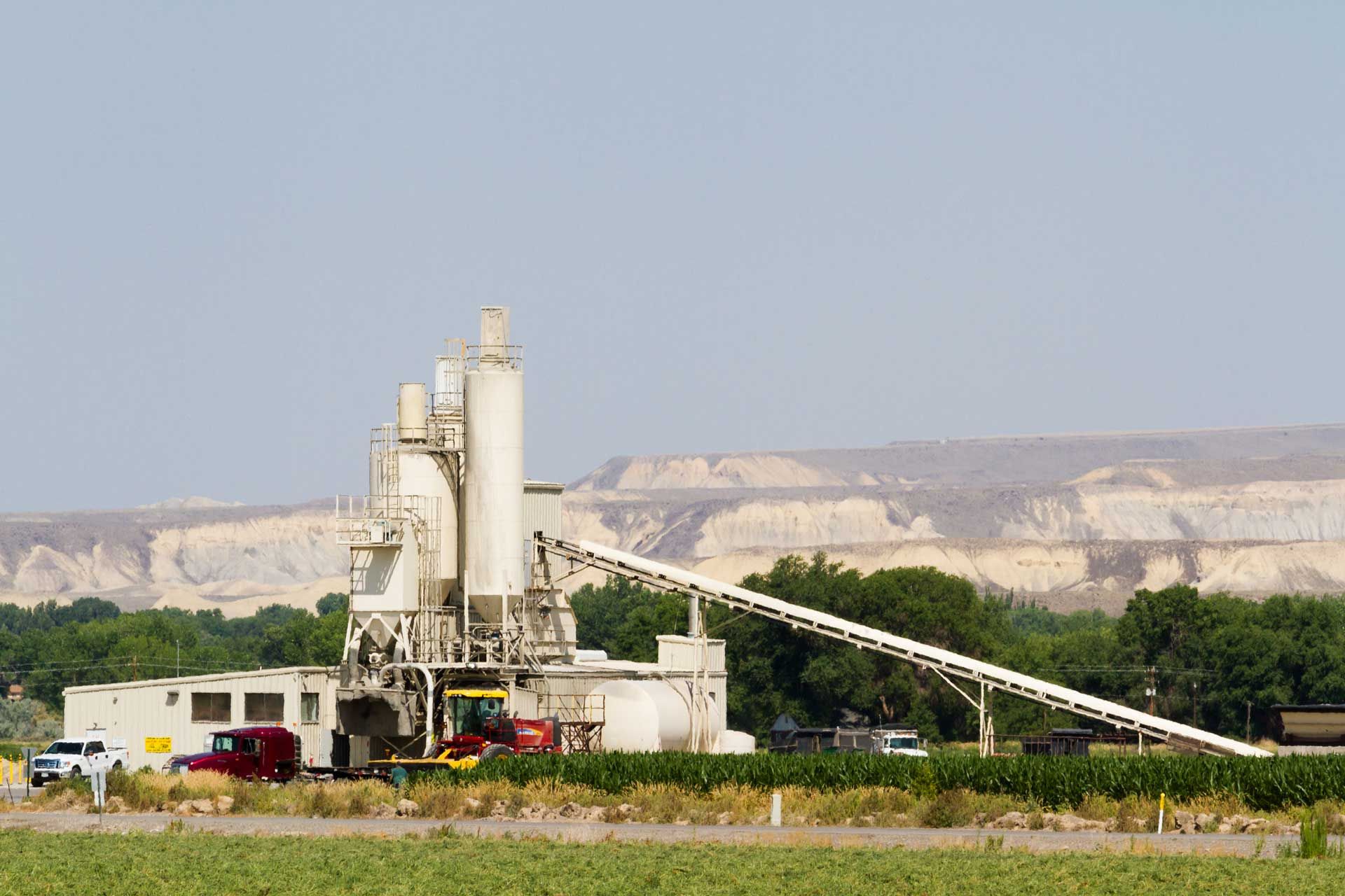 Cement plant with conveyor belt, red truck, and beige backdrop of hills under a blue sky.