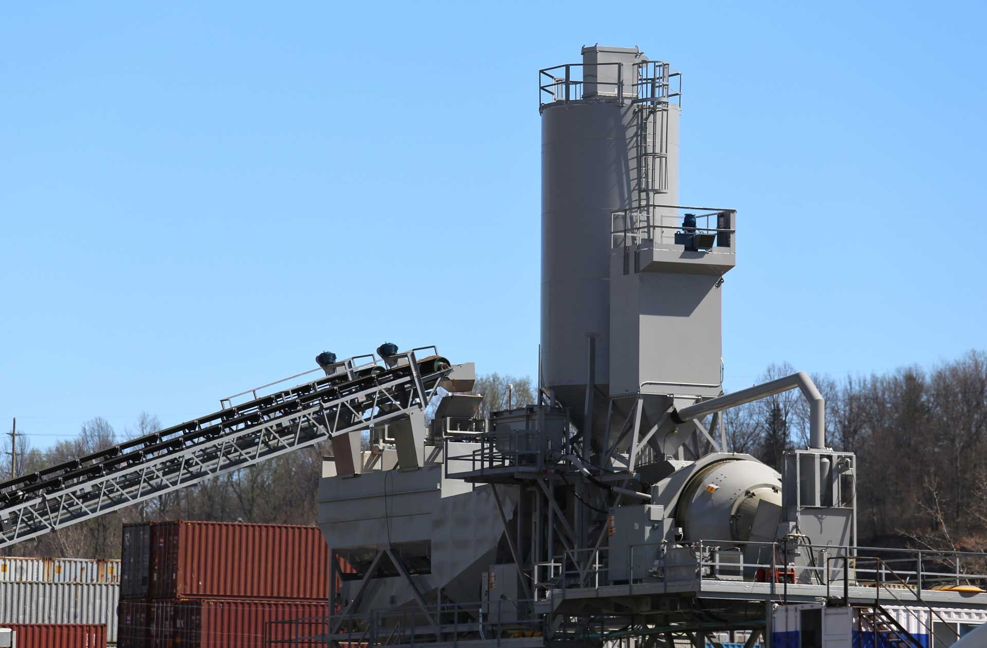 Concrete batch plant with conveyor belt, silo tower, and a mixer, under a blue sky.