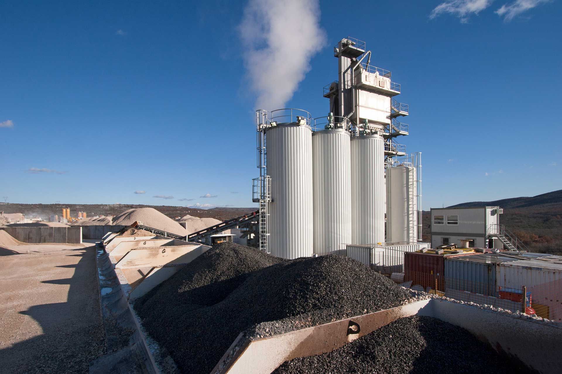 Asphalt plant with silos, steam, and piles of black asphalt under a blue sky.