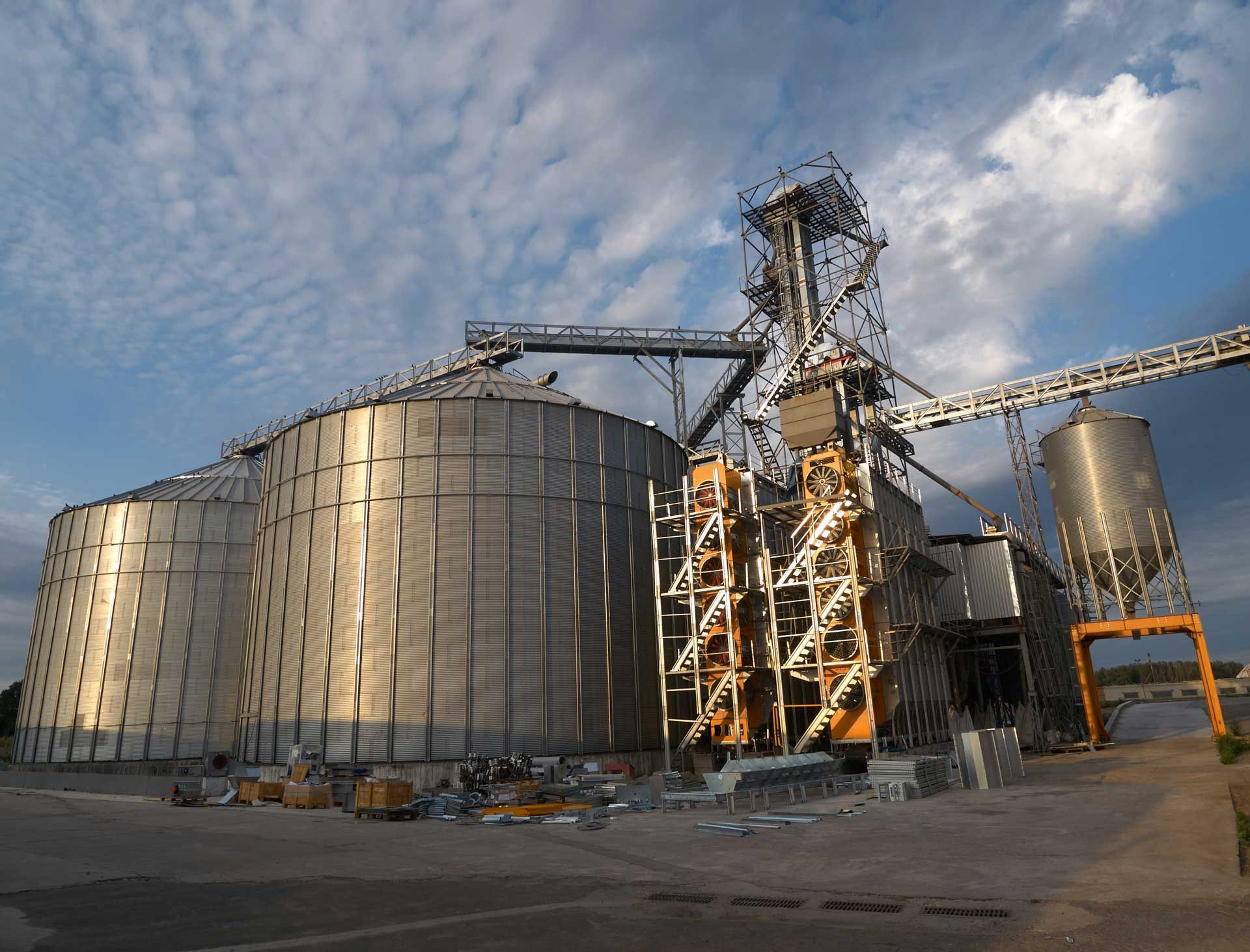 Grain storage facility with large silos and conveyor system against a cloudy sky.