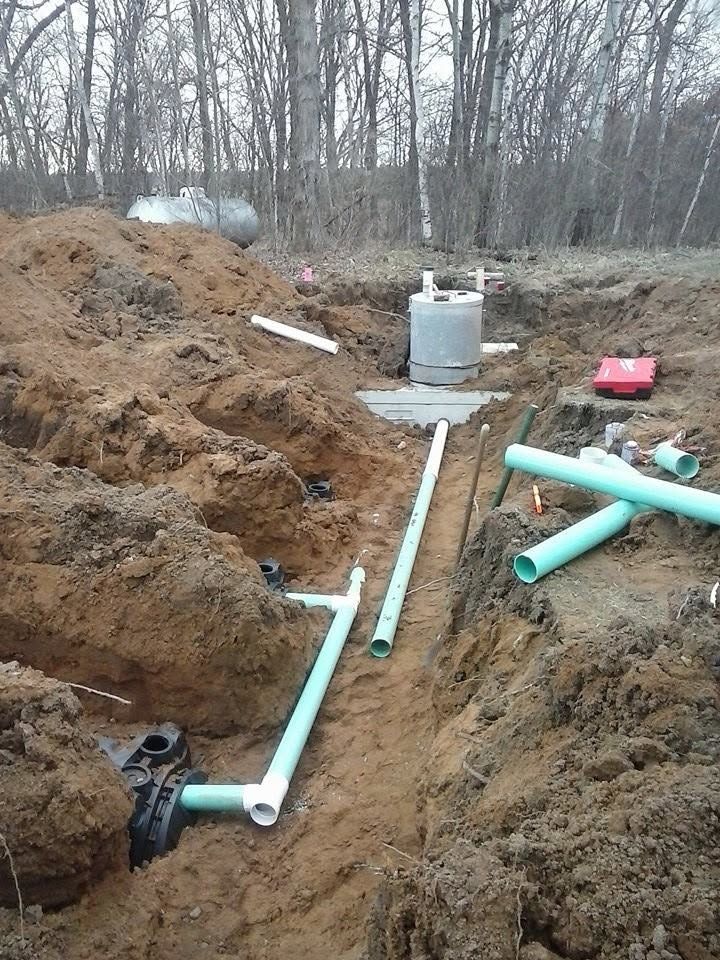 Trenched PVC pipes laid in a muddy wooded construction site with a septic tank and tools nearby