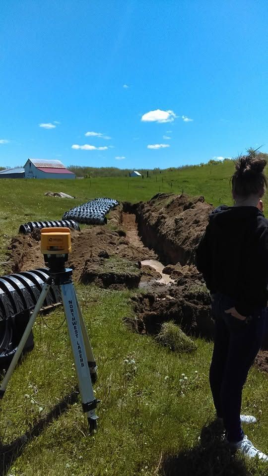 Person beside a digging trench in a sunny field, with surveying equipment and a distant barn under blue sky