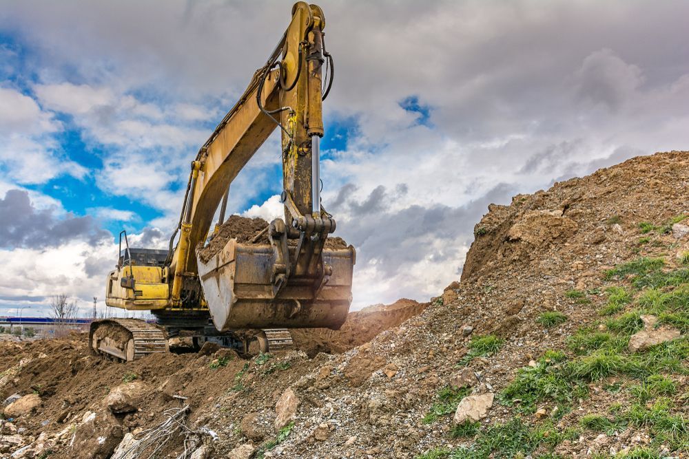 Yellow Excavator Digging Beside a Dirt Mound Under a Cloudy Sky — GPE Plumbers In Kendall, NSW