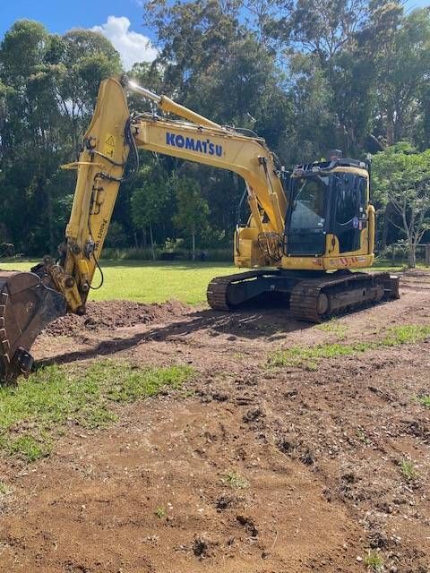A Yellow Excavator Operates on a Dirt Mound in Front of Two Tall — GPE Plumbers In Kendall, NSW