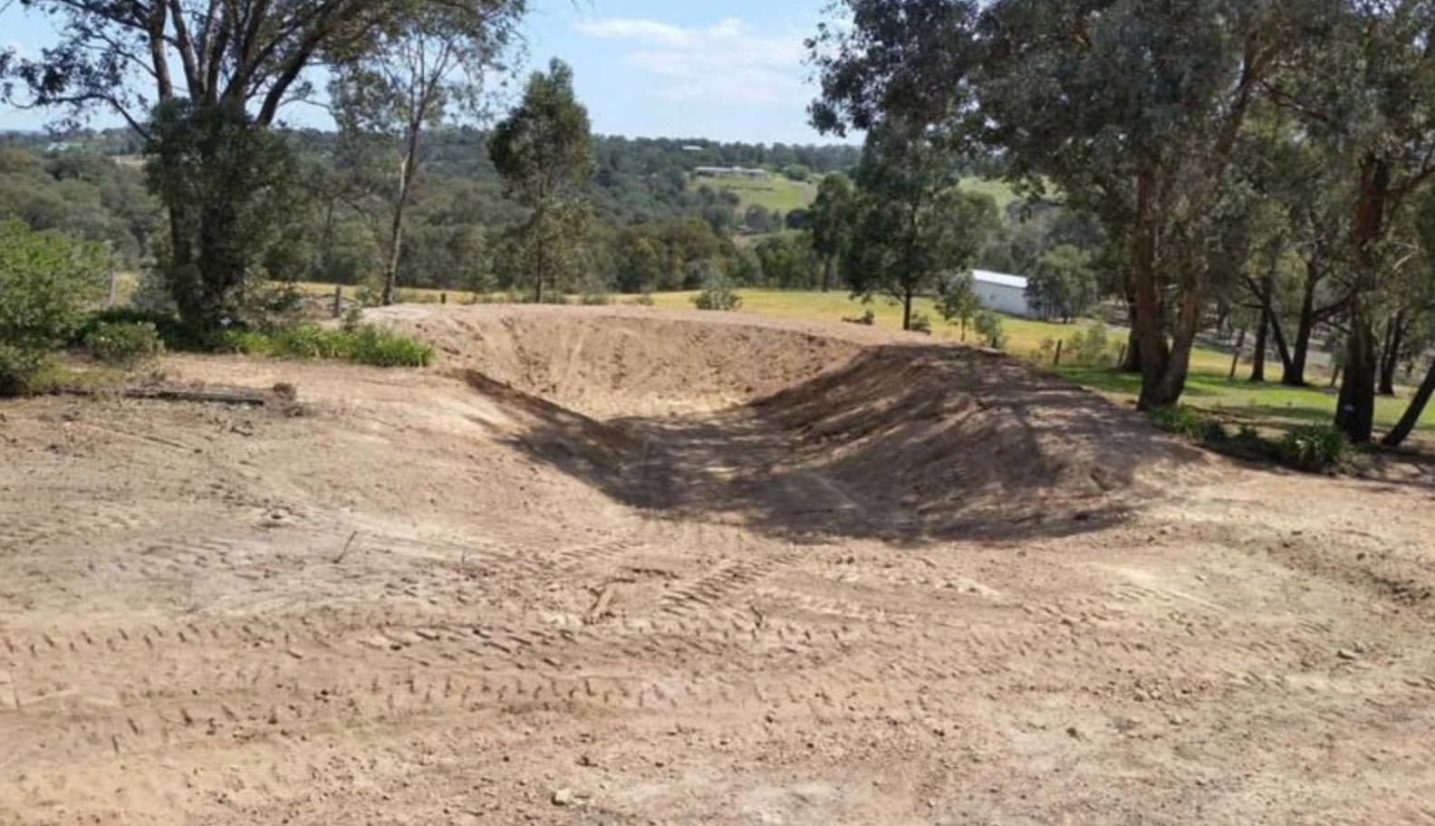 A newly excavated earth basin or pond in a grassy, wooded rural landscape under a clear blue sky — GPE Plumbers In Kendall, NSW