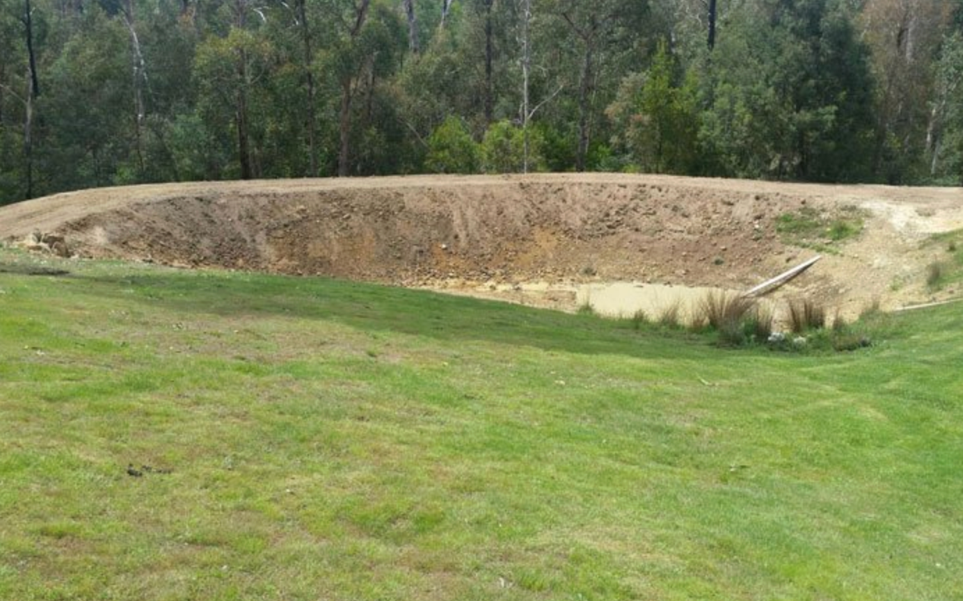A large, circular earthen pit dug into a grassy field in front of a forest line — GPE Plumbers In Kendall, NSW