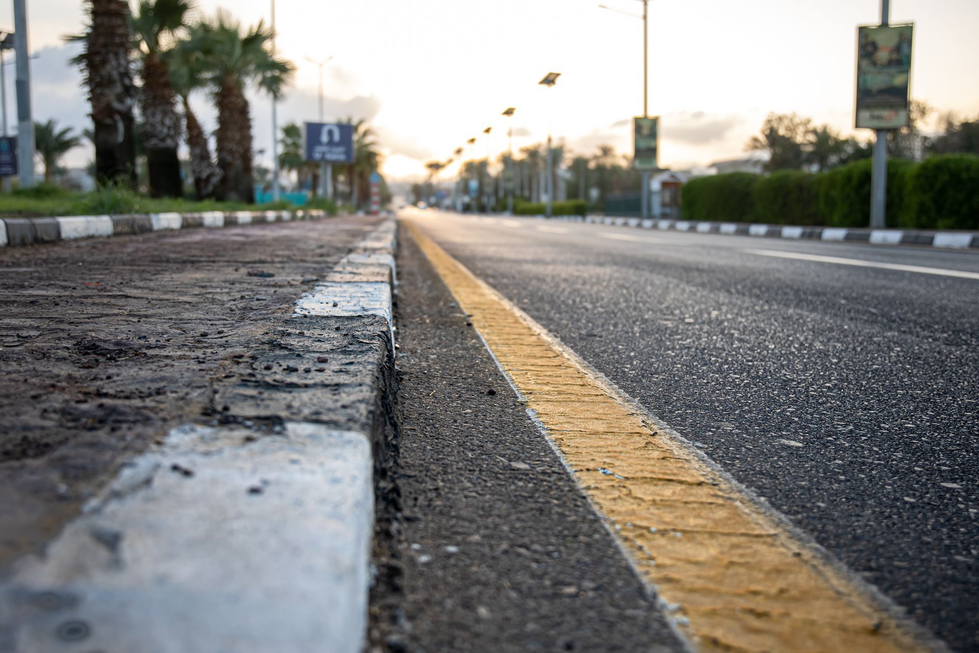 Low-angle view of a paved road with a yellow line and a black-and-white curb, alongside palm trees at sunset  — GPE Plumbers In Kendall, NSW