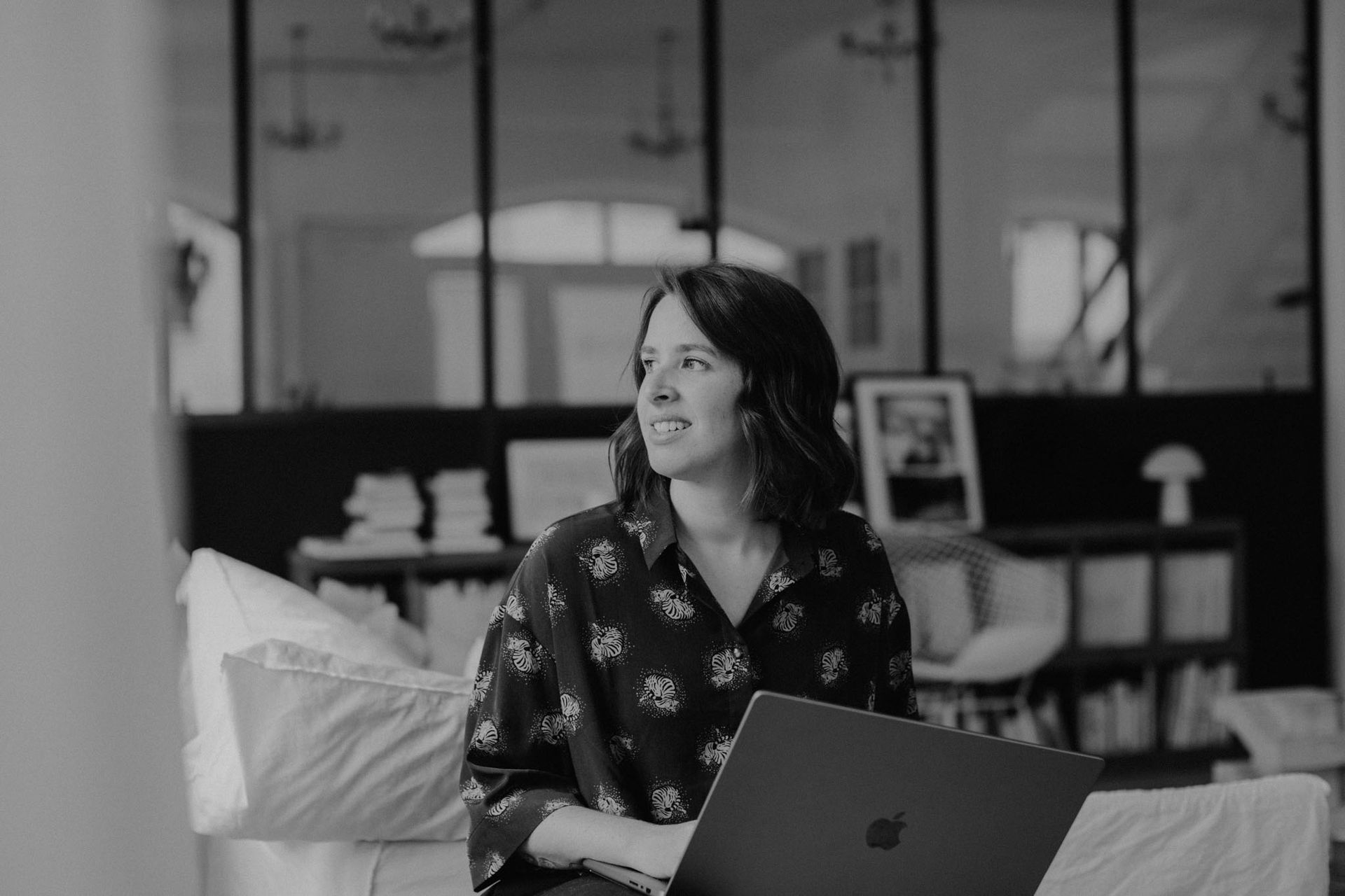 Sabrina mit Laptop, lächelt, schaut weg. Drinnen, sitzt auf einem Sofa, dunkles Hemd mit Tigern drauf.