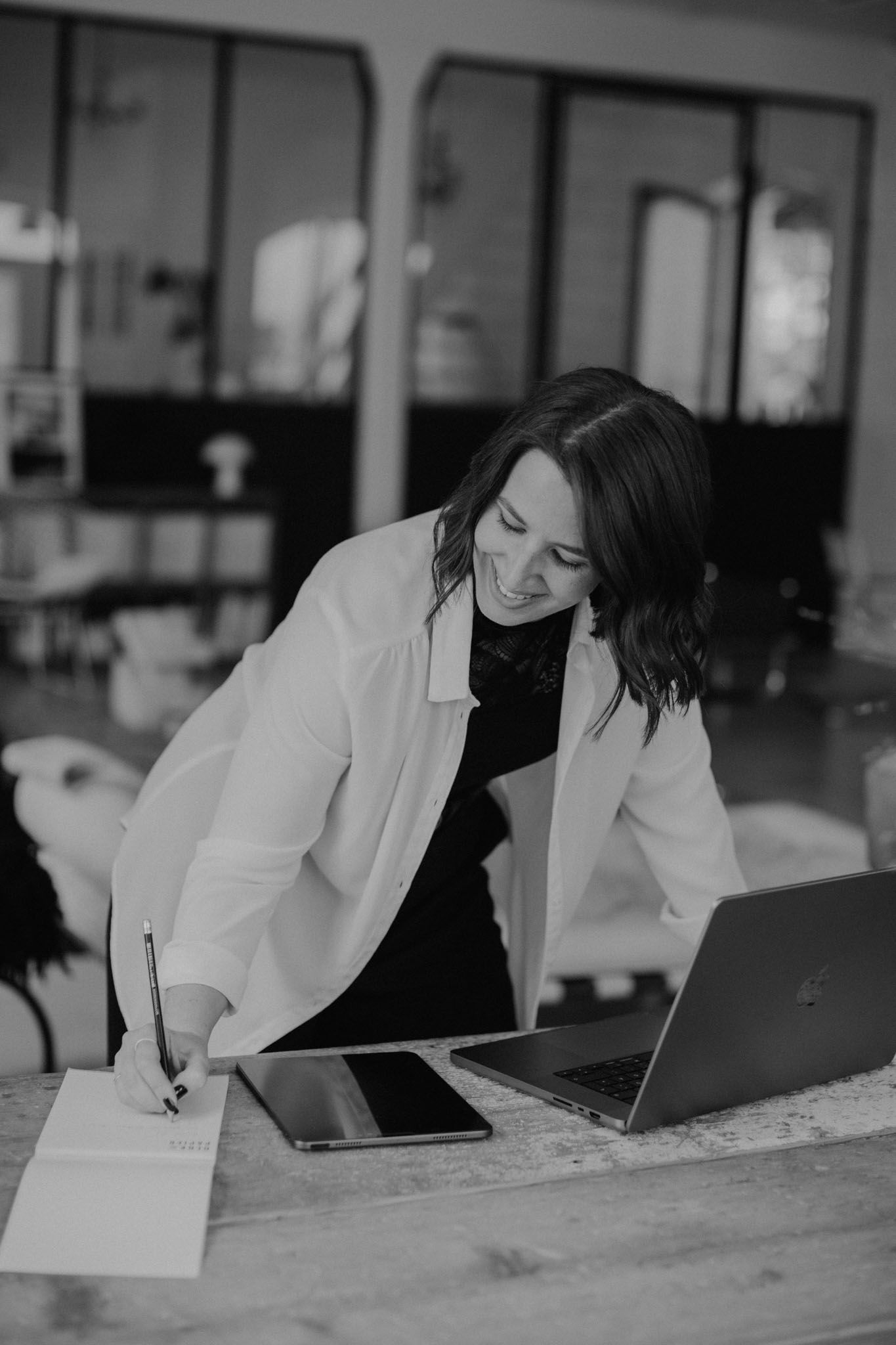 Sabrina lächelt, beugt sich über den Schreibtisch, schreibt in einen Notizblock; Laptop und Tablet sind vorhanden. Innenraum.