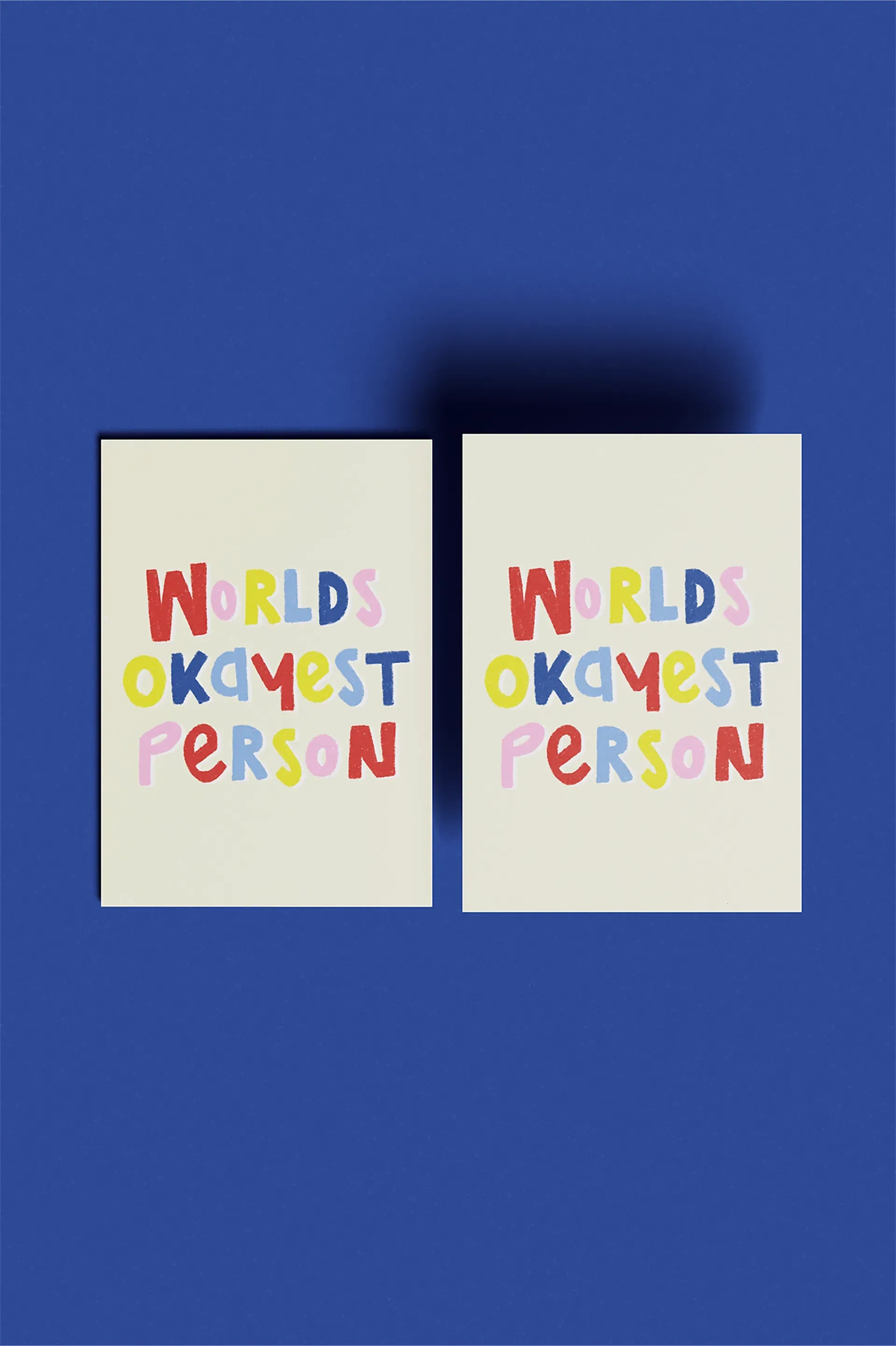 Zwei Postkarten mit der Aufschrift „Worlds Okayest Person“ in bunten Buchstaben auf blauem Hintergrund.