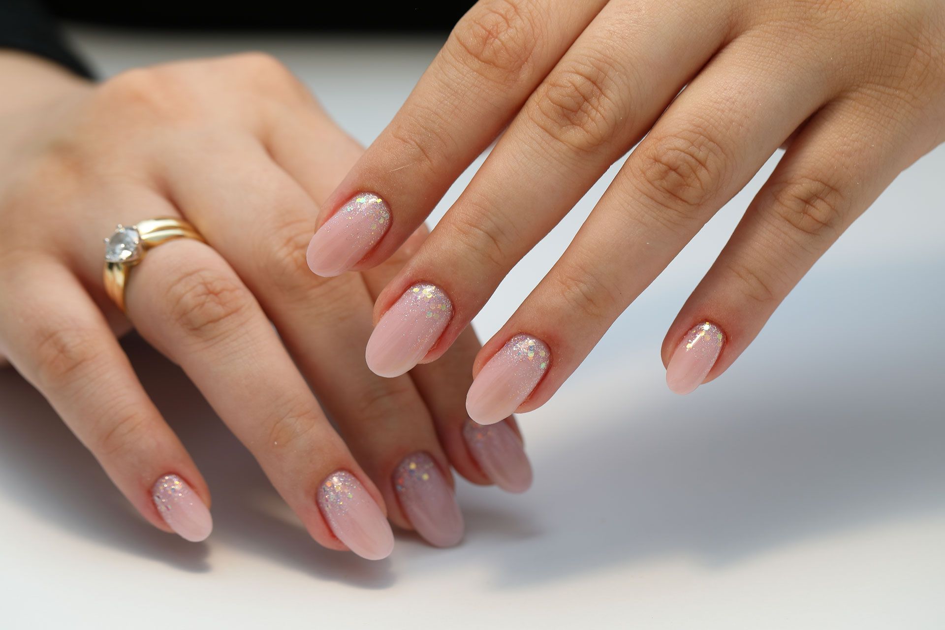 Hands showing off a professional manicure with light pink polish and subtle glitter accents, wearing a gold ring.